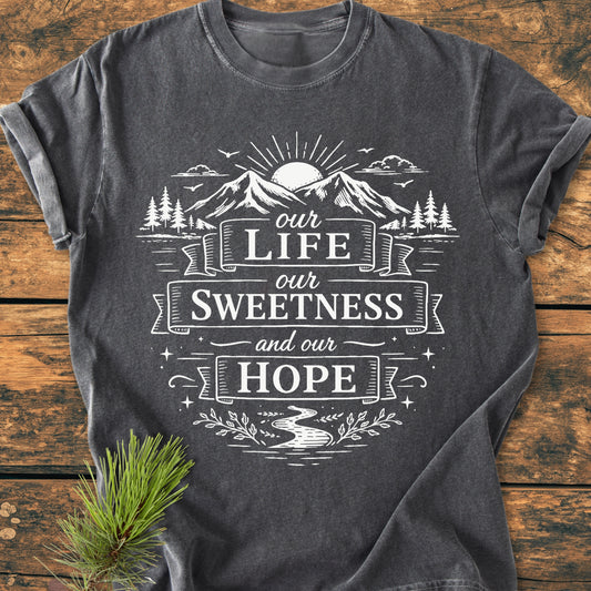 Our Life, Our Sweetness, Our Hope - Vintage Wash Tee features a stylized outdoor scene with sun, mountains, and river, printed on a charcoal-gray shirt, styled with a pine branch beside.