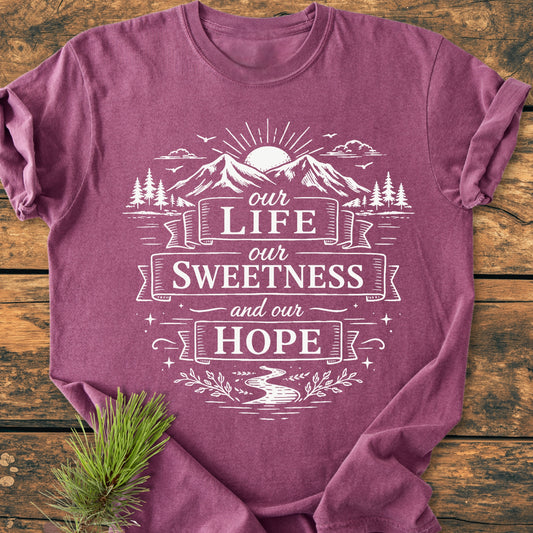 Our Life, Our Sweetness, Our Hope - Vintage Wash Tee featuring a mountain-and-sun graphic with trees, birds, and a stream, laid flat on wood with a pine branch accent.