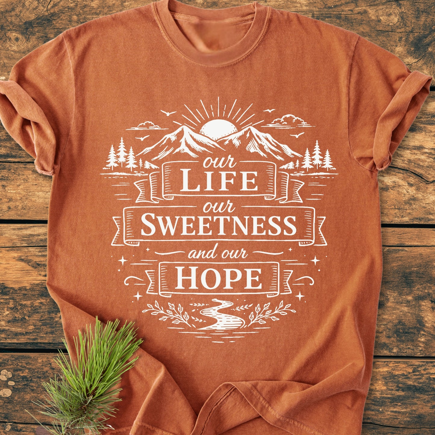 The Our Life, Our Sweetness, Our Hope - Vintage Wash Tee features a mountain landscape screen-printed on a burnt-orange shirt, emphasizing comfort and style, laid on a wooden surface with a pine sprig accent.