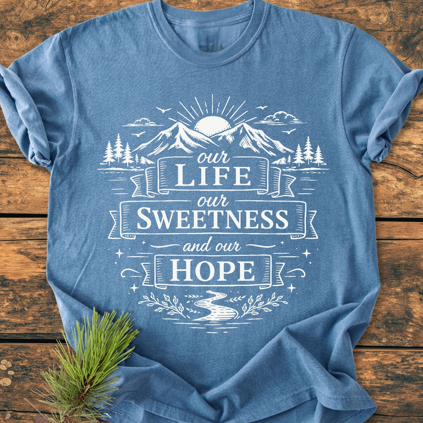 Our Life, Our Sweetness, Our Hope - Vintage Wash Tee features a nature-themed design with mountains, sunrise, pine trees, and banners. Laid on wood, complemented by a pine sprig for a rustic touch.