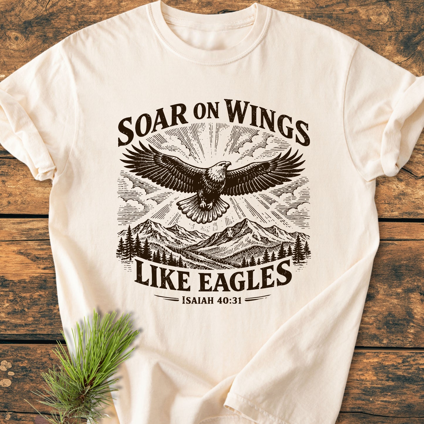 Soar On Wings Like Eagles Isaiah 40:31 - Vintage Wash Tee featuring an eagle illustration over mountains and sun rays, laid flat with a pine sprig, showcasing rustic, inspirational design.