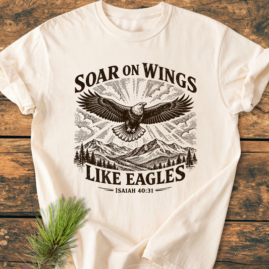 Soar On Wings Like Eagles Isaiah 40:31 - Vintage Wash Tee featuring an eagle illustration over mountains and sun rays, laid flat with a pine sprig, showcasing rustic, inspirational design.