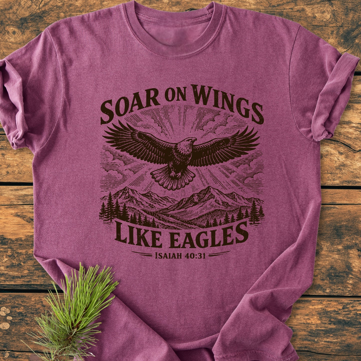 Soar On Wings Like Eagles Isaiah 40:31 - Vintage Wash Tee: Heather-purple T-shirt with stylized eagle, mountain, and forest print; pine sprig beside, laid on wooden surface, highlighting comfort and relaxed fit.