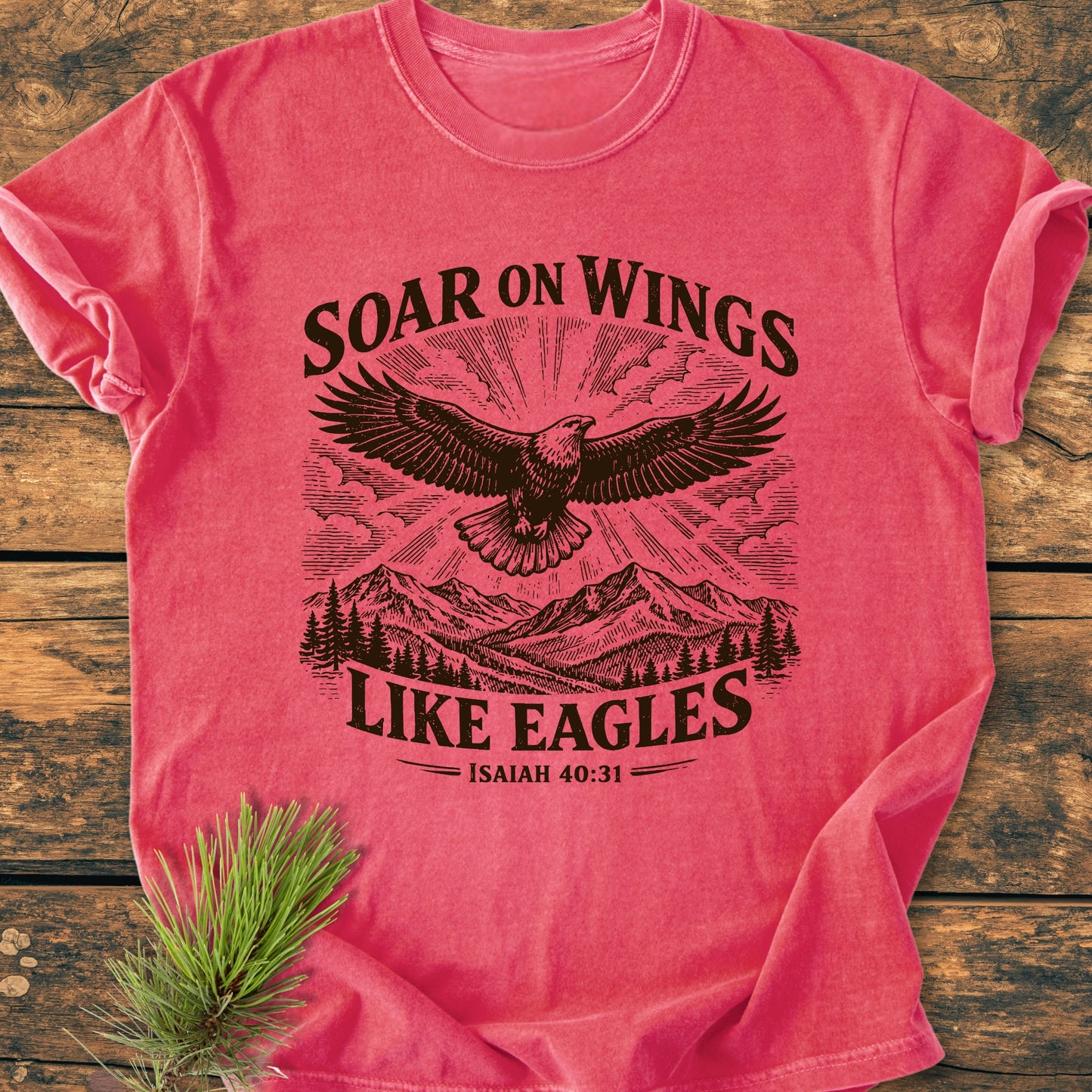 Soar On Wings Like Eagles Isaiah 40:31 - Vintage Wash Tee features an eagle graphic with pine trees and mountains, on coral-pink fabric, laid on wood, with a pine sprig beside it.