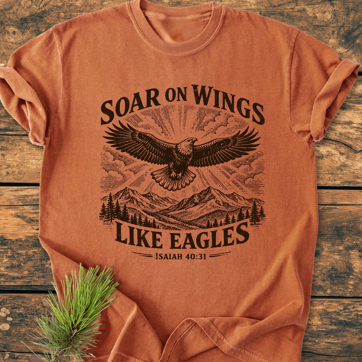 Soar On Wings Like Eagles Isaiah 40:31 - Vintage Wash Tee features a stylized eagle graphic over mountains and pine trees on a rust-orange T-shirt, laid flat on wooden surface.