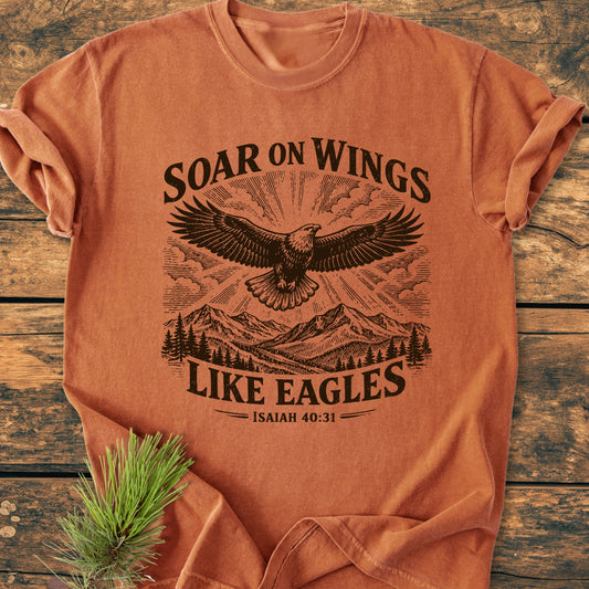 Soar On Wings Like Eagles Isaiah 40:31 - Vintage Wash Tee features a stylized eagle graphic over mountains and pine trees on a rust-orange T-shirt, laid flat on wooden surface.