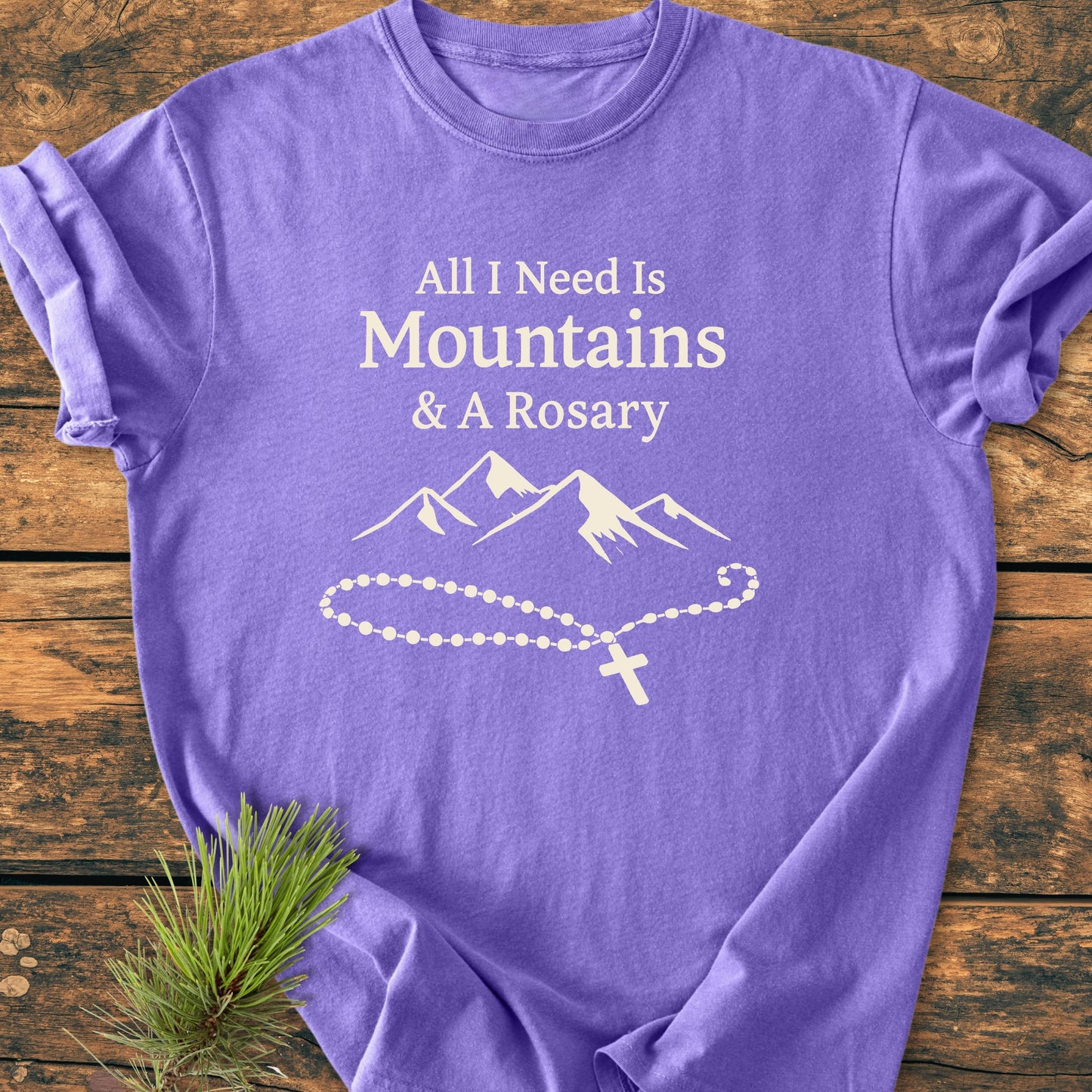 Laid-flat All I Need Is Mountains And A Rosary - Vintage Wash Tee with white text, mountain drawing, and rosary on purple fabric, displayed on wood, featuring a soft, relaxed fit and durable stitching.
