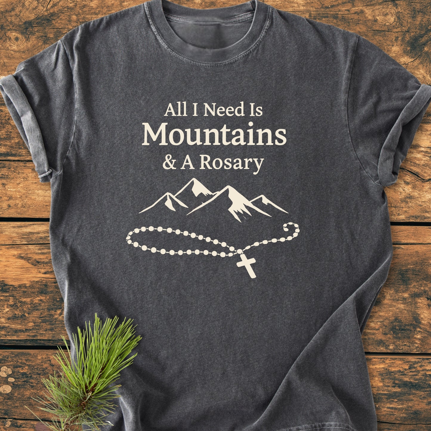 Charcoal-gray All I Need Is Mountains And A Rosary - Vintage Wash Tee, featuring white mountain and rosary print, laid on wood with pine sprig. Soft, relaxed fit, 100% ring-spun cotton.