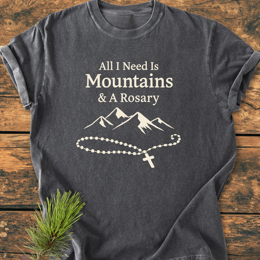 Charcoal-gray All I Need Is Mountains And A Rosary - Vintage Wash Tee, featuring white mountain and rosary print, laid on wood with pine sprig. Soft, relaxed fit, 100% ring-spun cotton.
