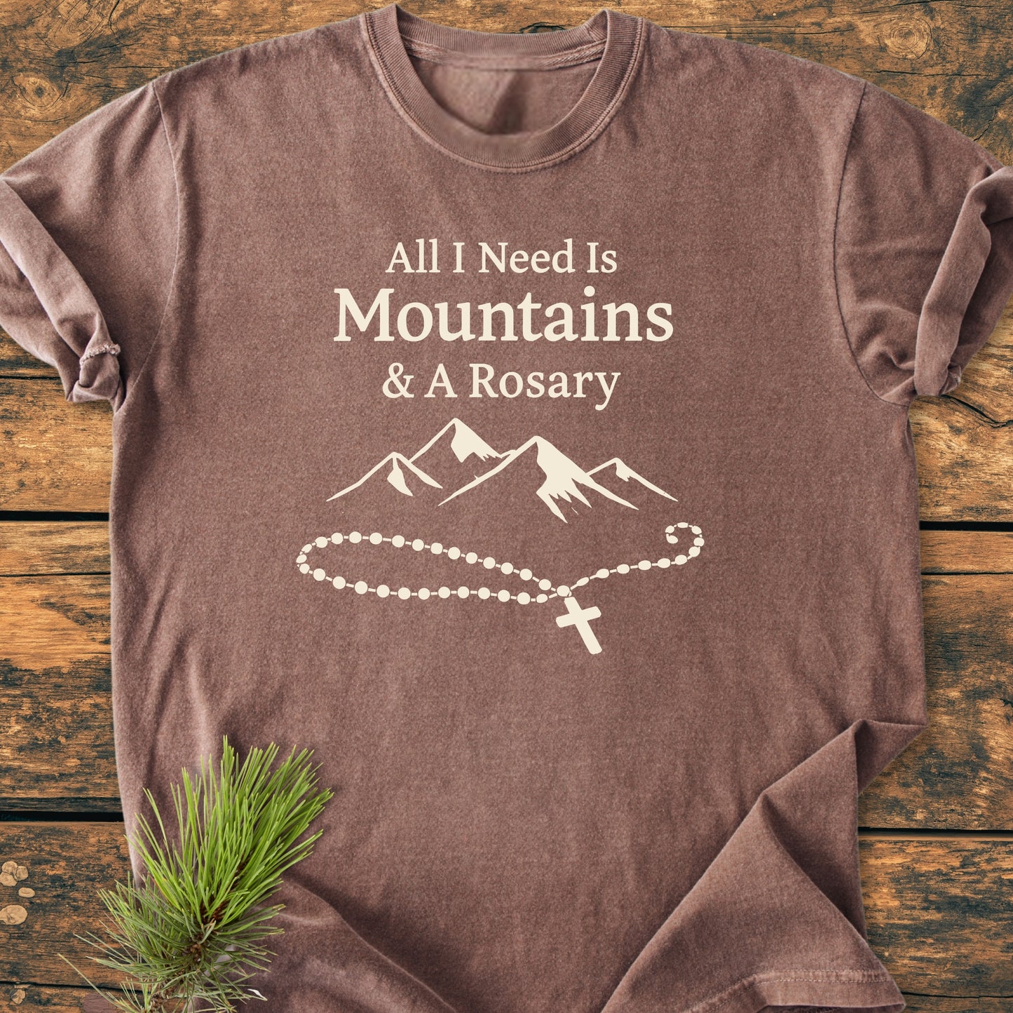 All I Need Is Mountains And A Rosary - Vintage Wash Tee displayed on wood, featuring a cream print of text, mountain range, and rosary, highlighting its soft, relaxed fit and durable design.