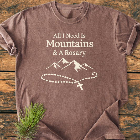 All I Need Is Mountains And A Rosary - Vintage Wash Tee displayed on wood, featuring a cream print of text, mountain range, and rosary, highlighting its soft, relaxed fit and durable design.