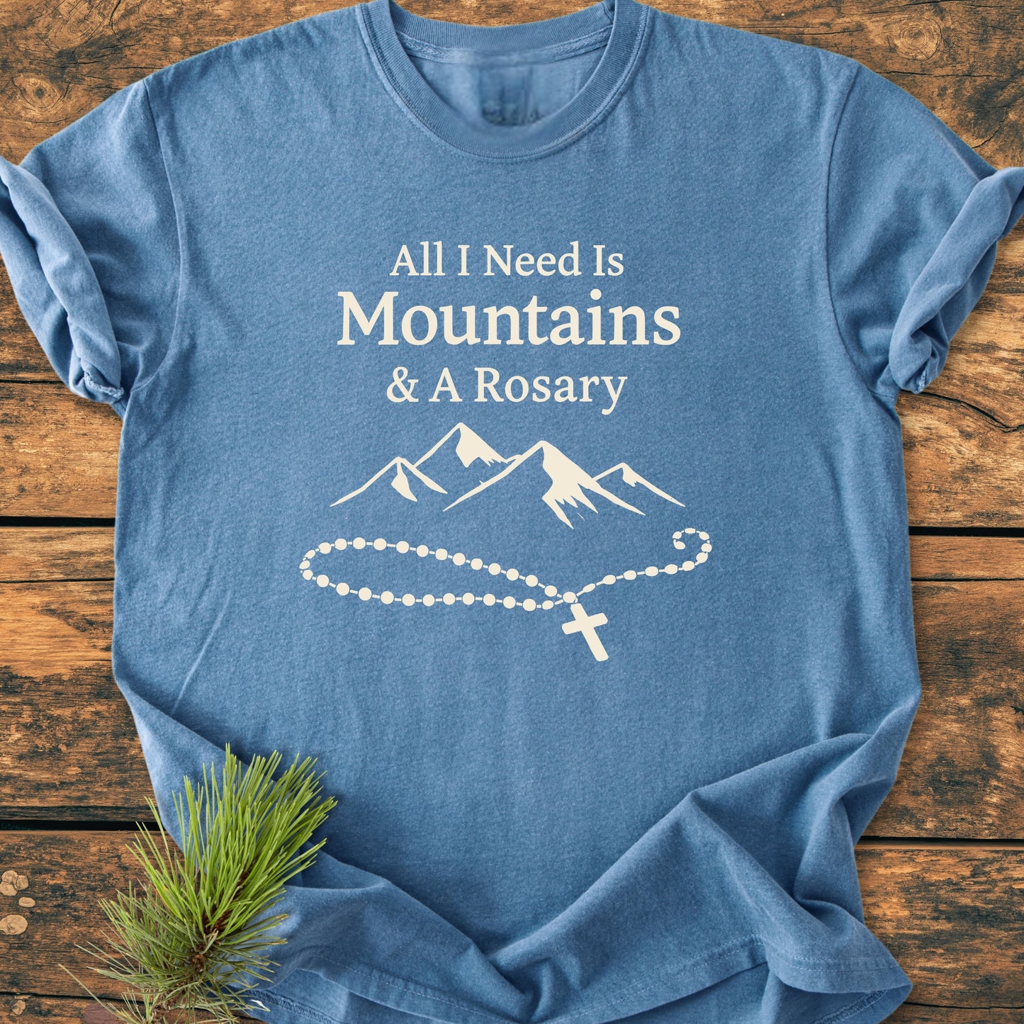 Heather-blue “All I Need Is Mountains And A Rosary - Vintage Wash Tee” with white text and mountain-rosary graphic, laid flat on wood, showcasing its relaxed fit and durable, soft-washed fabric.