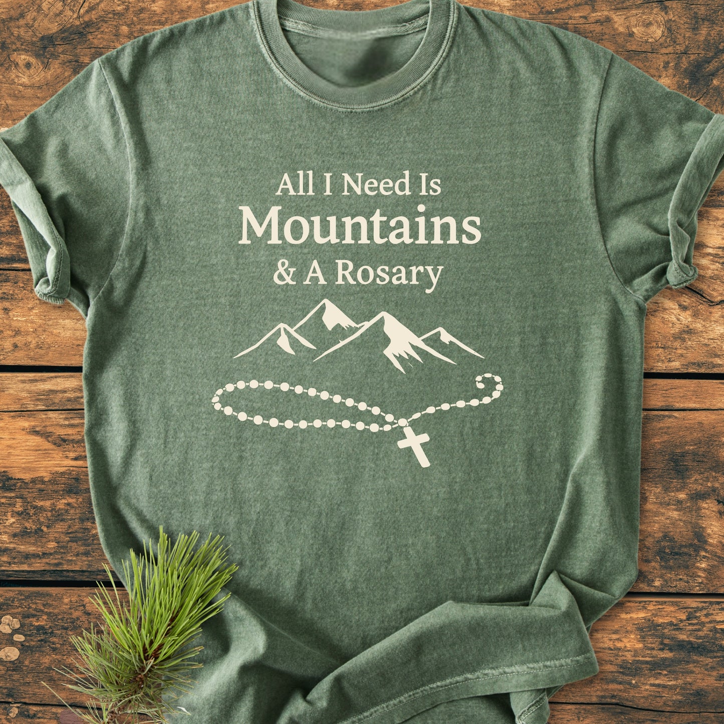 Forest-green vintage wash tee with “All I Need Is Mountains & A Rosary” in off-white, featuring a mountain graphic and rosary outline, laid on wood with a pine sprig beside it.