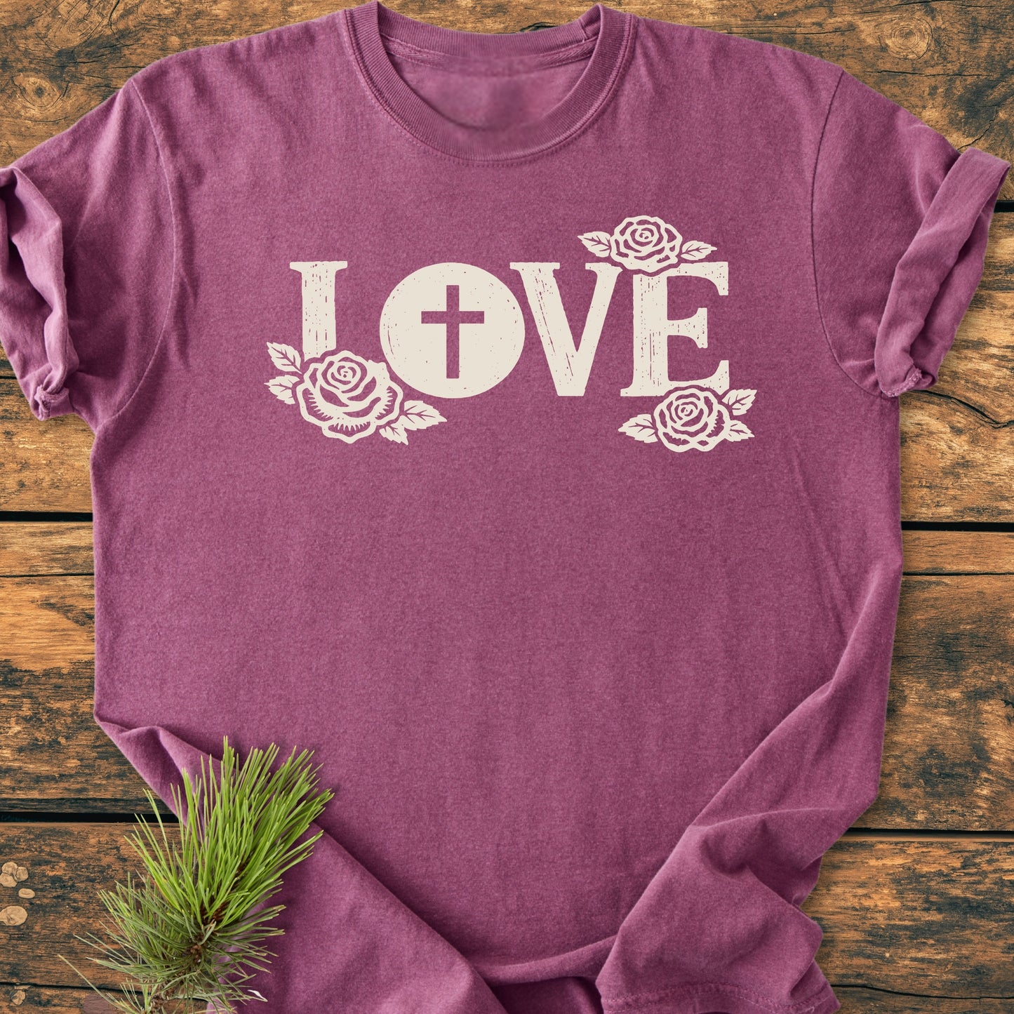 The LOVE – Eucharistic Faith - Vintage Wash Tee features LOVE with a cross in the O and rose accents, laid flat on wood. Includes pine sprig detail.