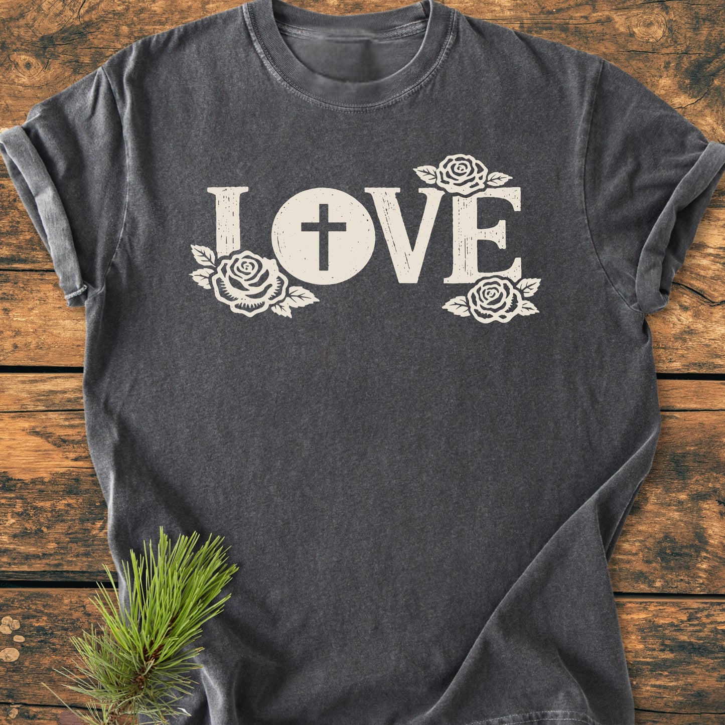 LOVE – Eucharistic Faith - Vintage Wash Tee features “LOVE” in distressed font with rose accents on a charcoal-gray T-shirt, laid flat on rustic wood, highlighting its soft, garment-dyed cotton fabric.