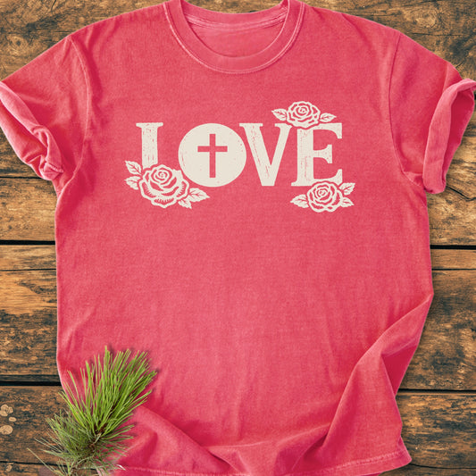 LOVE – Eucharistic Faith - Vintage Wash Tee, featuring a distressed white “LOVE” print with a cross and roses, laid flat on wood, soft-washed for comfort with a relaxed fit.