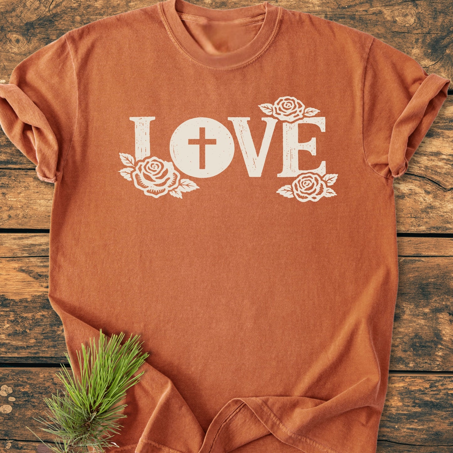 LOVE – Eucharistic Faith - Vintage Wash Tee: Rust-orange T-shirt featuring LOVE with a cross in the O and white roses; laid on wood with a pine sprig accent.