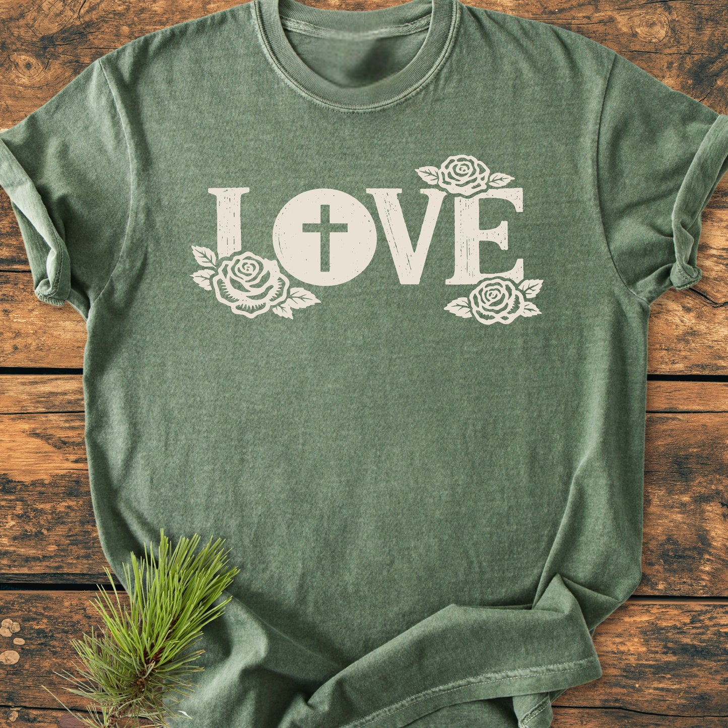 LOVE – Eucharistic Faith - Vintage Wash Tee featuring a forest-green color, short sleeves, LOVE with a cross and rose accents, laid on a wooden surface with a pine sprig nearby.
