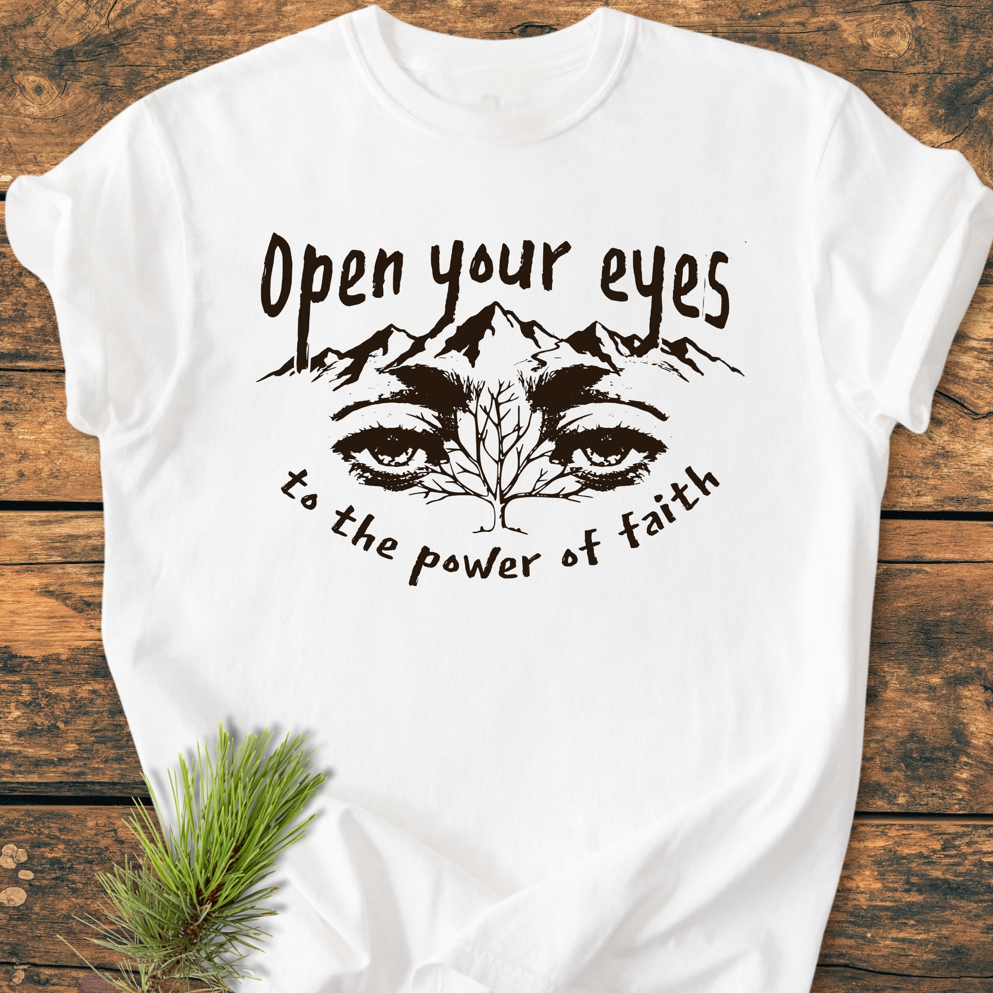 Open Your Eyes To The Power Of Faith - Vintage Wash Tee featuring a mountain and eyes graphic on soft, garment-dyed cotton, displayed with a pine sprig on a wooden surface.