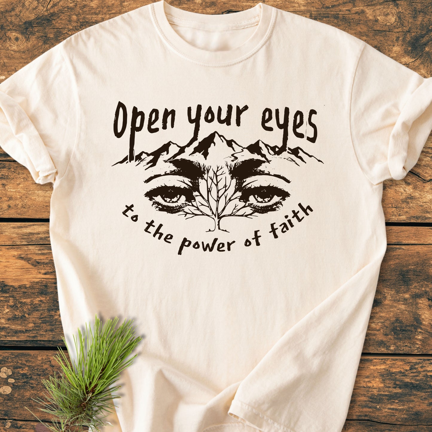Open Your Eyes To The Power Of Faith - Vintage Wash Tee; off-white cotton with mountain-eye graphic and phrase, laid on rustic wood with pine sprig, showcasing relaxed fit and durable stitching.