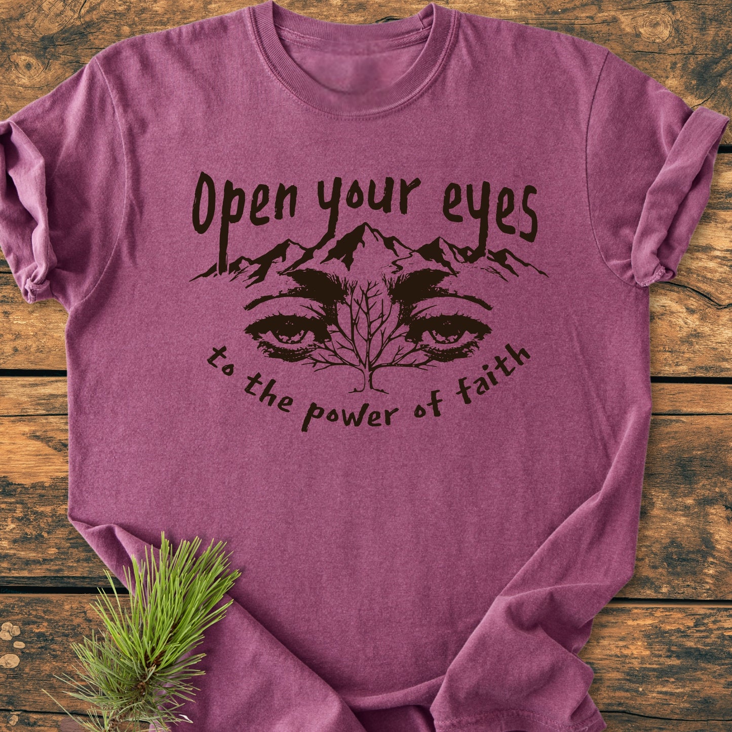 Open Your Eyes To The Power Of Faith - Vintage Wash Tee features a graphic of eyes and mountain branches, with inspiring text, laid on a wooden surface beside an evergreen sprig.