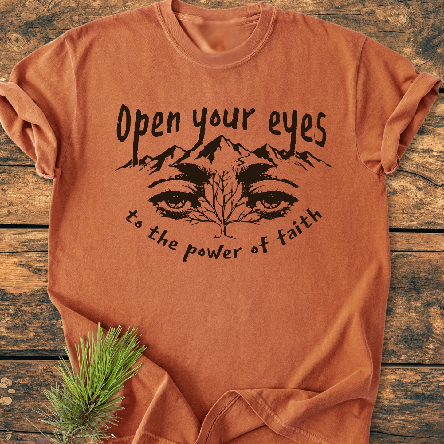 Open Your Eyes To The Power Of Faith - Vintage Wash Tee features a mountain and tree design with inspirational text, displayed on a rust-orange t-shirt, accompanied by a small evergreen sprig.