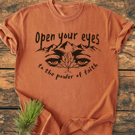 Open Your Eyes To The Power Of Faith - Vintage Wash Tee features a mountain and tree design with inspirational text, displayed on a rust-orange t-shirt, accompanied by a small evergreen sprig.