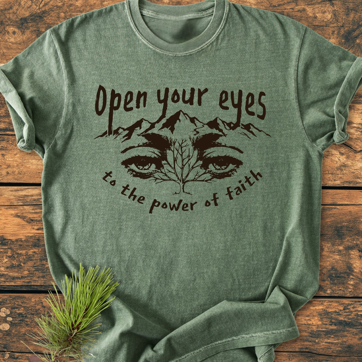 Open Your Eyes To The Power Of Faith - Vintage Wash Tee features a nature graphic with mountain peaks and eyes on green fabric, laid on wood with a pine sprig for a rustic look.