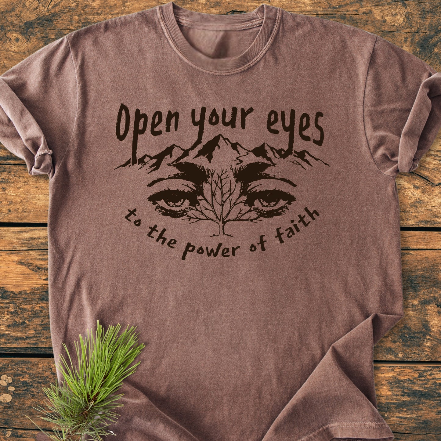 Open Your Eyes To The Power Of Faith - Vintage Wash Tee laid flat, featuring a mountain graphic, tree, eyes, and slogan, with a pine sprig accent on a rustic wooden background.