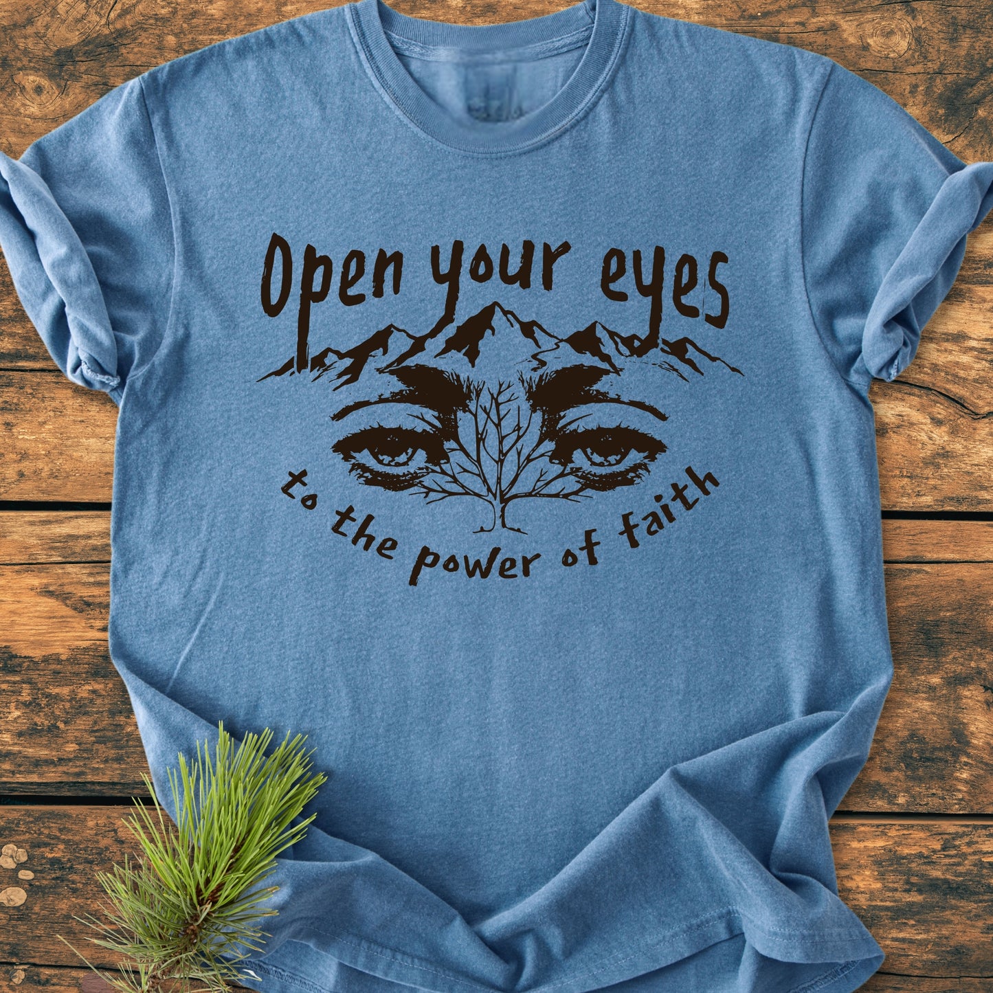 Open Your Eyes To The Power Of Faith - Vintage Wash Tee: Heather-blue T-shirt with a black graphic of eyes, mountains, and tree, featuring inspirational phrases. Laid on wood with a pine sprig accent.