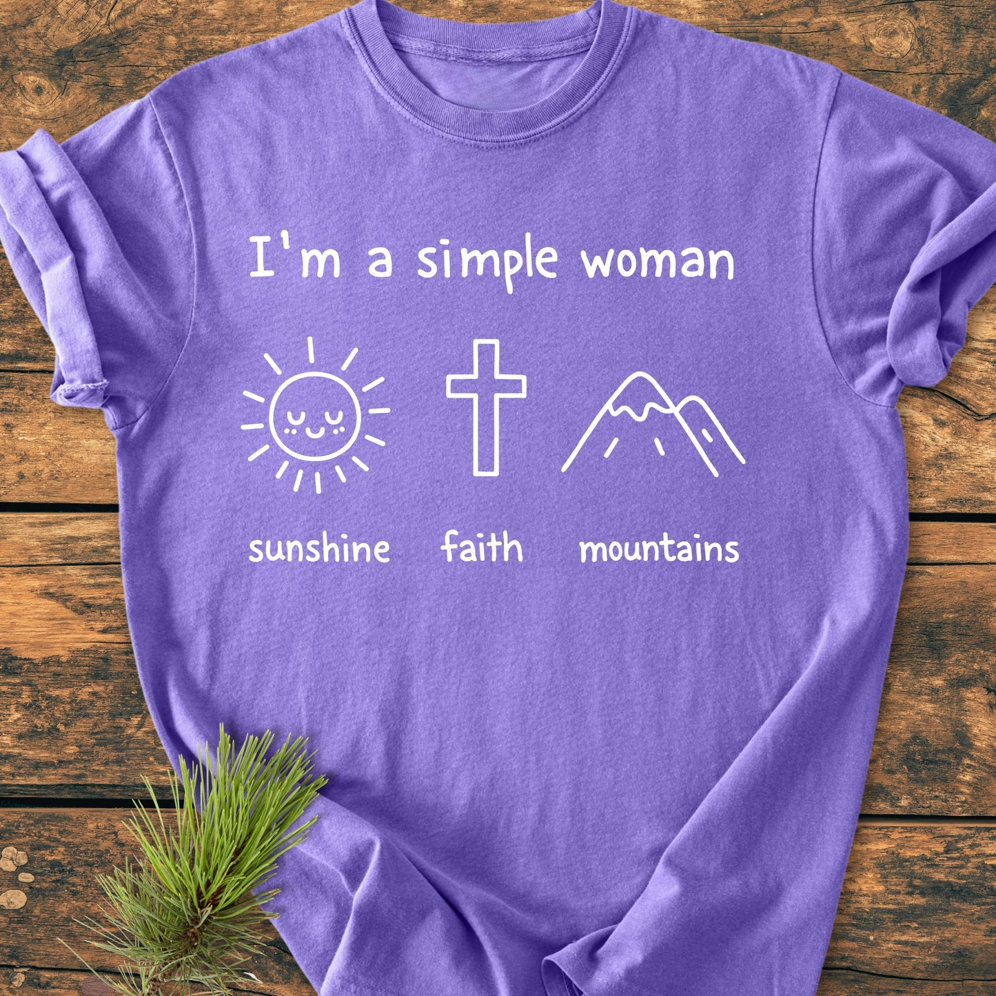 I'm A Simple Woman. Sunshine Faith Mountains - Vintage Wash Tee features white text and icons of a sun, cross, and mountains on a purple T-shirt, laid flat on a wood surface.