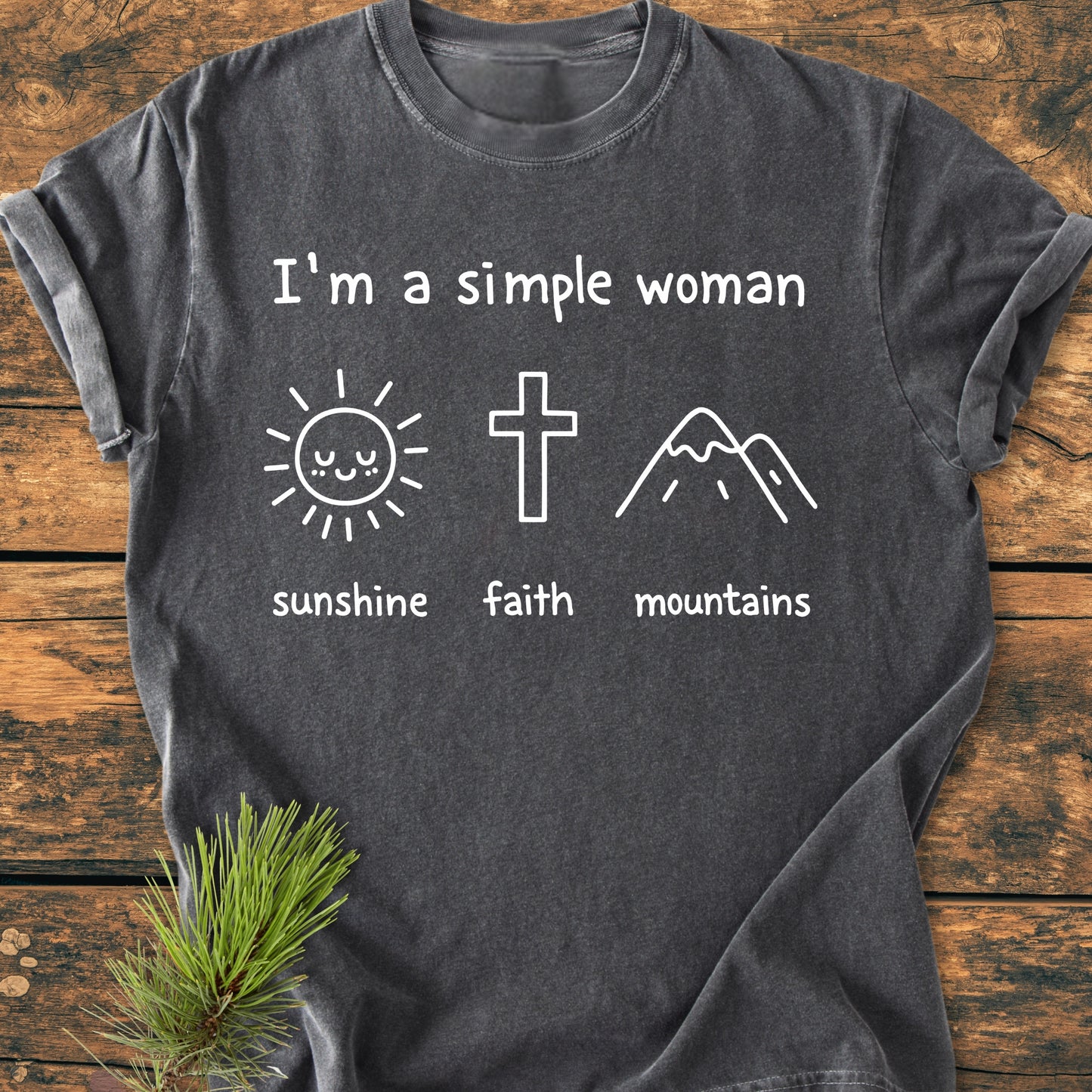 I'm A Simple Woman. Sunshine Faith Mountains - Vintage Wash Tee: A dark-gray T-shirt featuring white lettering and icons of a sun, cross, and mountains, laid flat on a wooden surface.