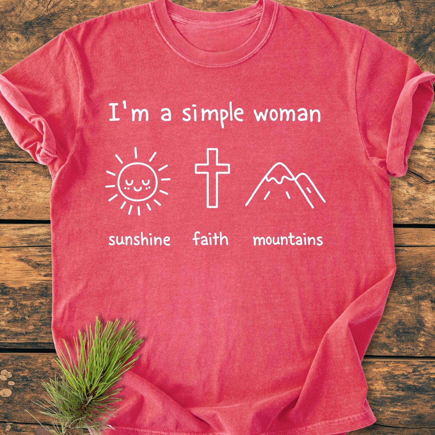 The I'm A Simple Woman. Sunshine Faith Mountains - Vintage Wash Tee features a coral-red color with white prints and icons, laid flat on a wooden surface, adorned by a small evergreen sprig.