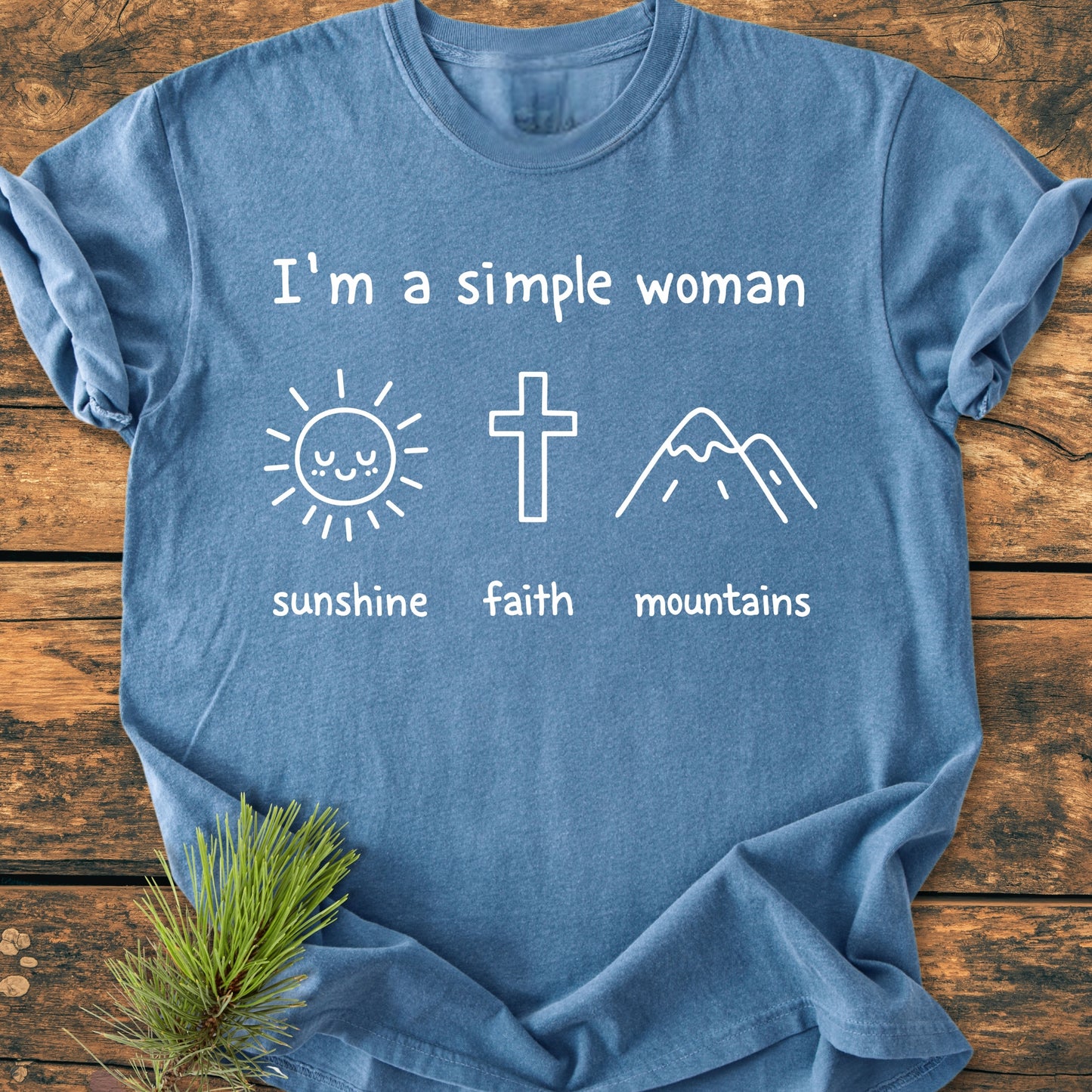 I'm A Simple Woman. Sunshine Faith Mountains - Vintage Wash Tee: Heather-blue T-shirt with white line art of a sun, cross, and mountains, laid flat on wood, pine sprig beneath corner.