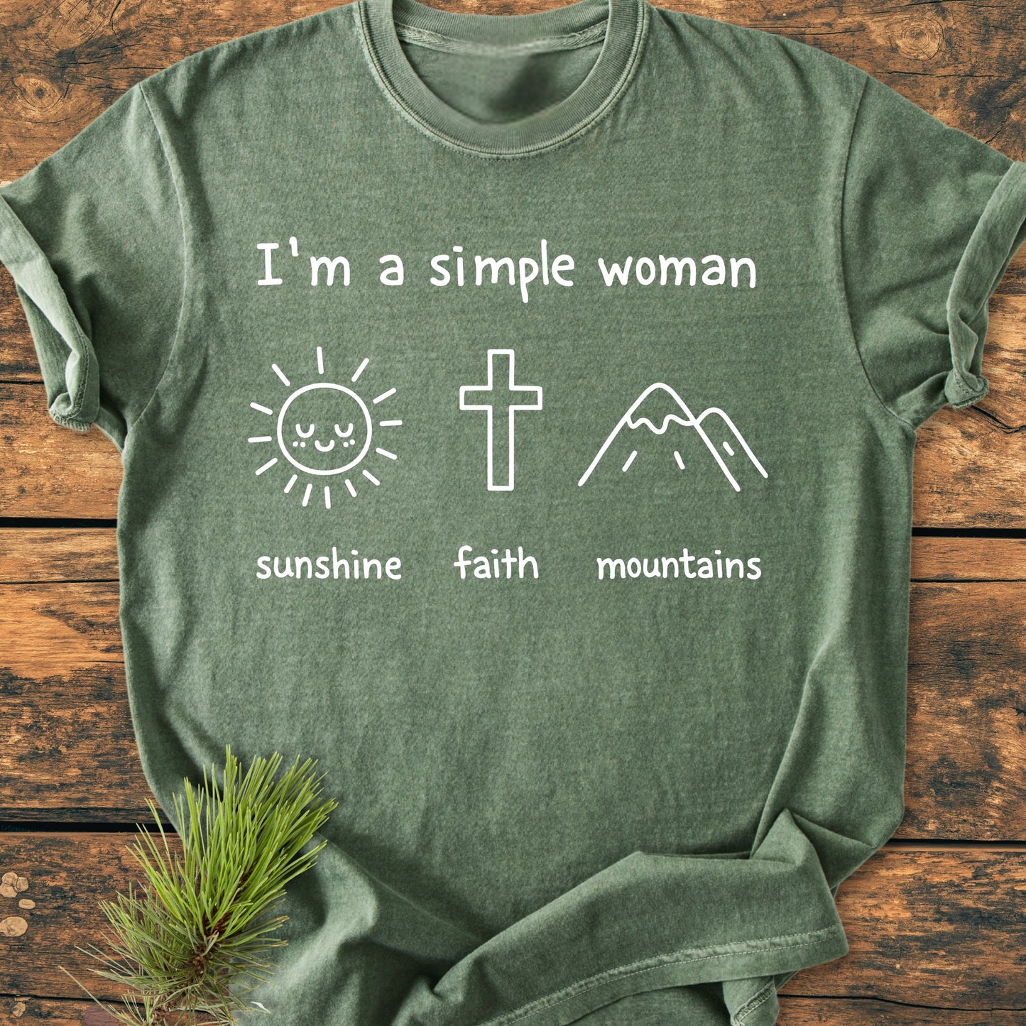 I'm A Simple Woman. Sunshine Faith Mountains - Vintage Wash Tee features a heather-green design with a sun, cross, and mountain icons, laid on wood, enhanced by a pine sprig.