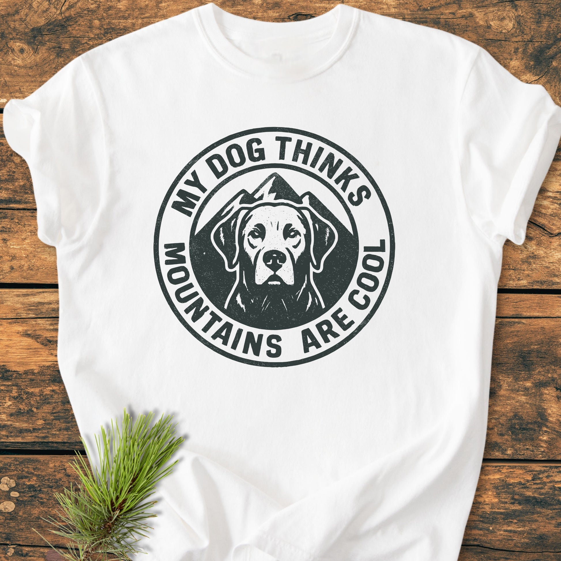 My Dog Thinks Mountains Are Cool - Vintage Wash Tee features a dog graphic with mountain silhouette on a white T-shirt, presented on a wooden surface with a pine sprig for natural touch.