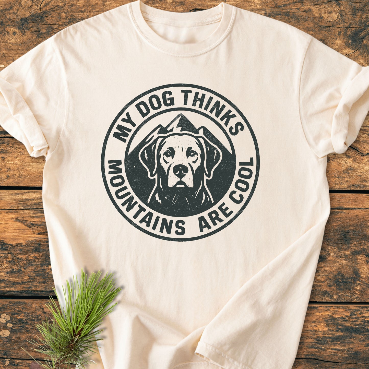Light-beige My Dog Thinks Mountains Are Cool - Vintage Wash Tee with a stamp-style graphic of a dog and mountains, laid flat on a wooden surface with a pine sprig nearby.