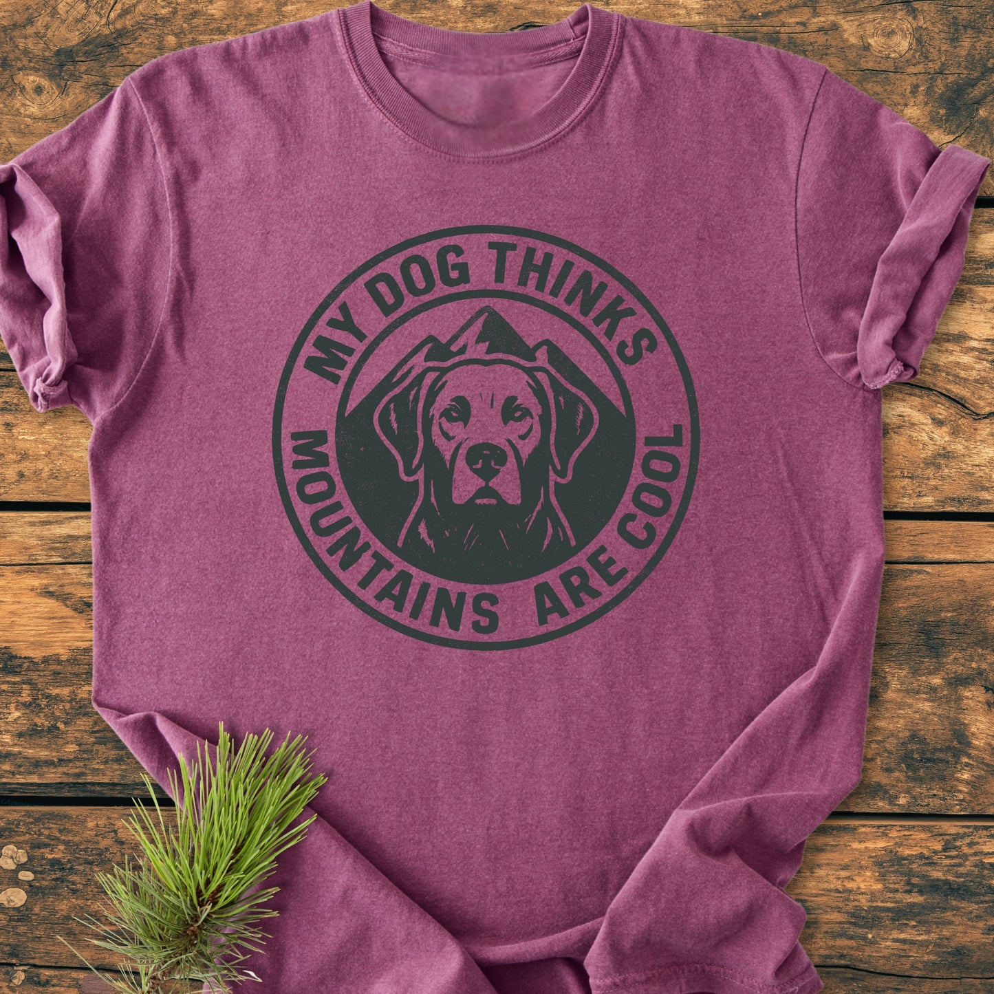 My Dog Thinks Mountains Are Cool - Vintage Wash Tee: Heather-purple crew-neck with black screen-printed badge showing a dog's head and mountain peaks, laid on a wooden surface with a pine sprig.