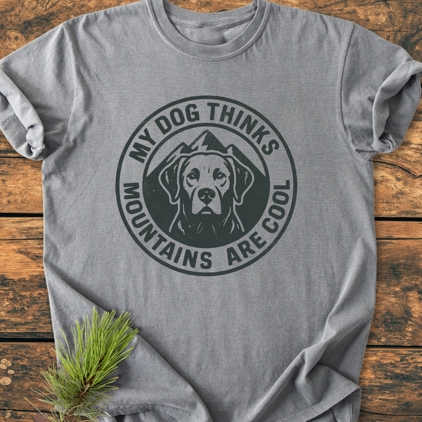 Heather-gray My Dog Thinks Mountains Are Cool - Vintage Wash Tee with circular dog and mountain graphic, laid flat on wood, pine branch nearby. Crafted from soft, pre-shrunk, ring-spun cotton for everyday comfort.