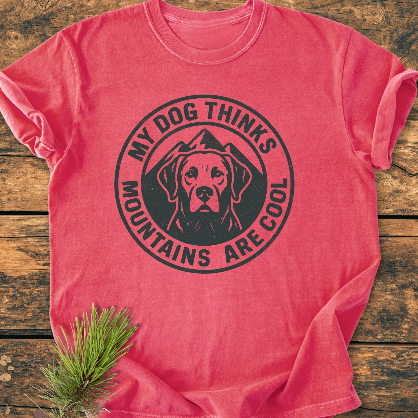 My Dog Thinks Mountains Are Cool - Vintage Wash Tee features a black dog graphic with mountains on a red shirt, laid on wood. Includes pine branch accent; soft, relaxed fit cotton.