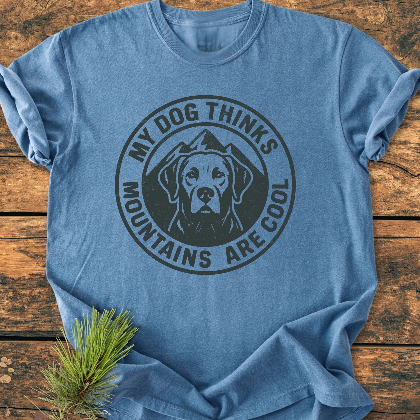 The My Dog Thinks Mountains Are Cool - Vintage Wash Tee features a black graphic of a dog with mountains, styled on a wooden surface with a pine sprig.