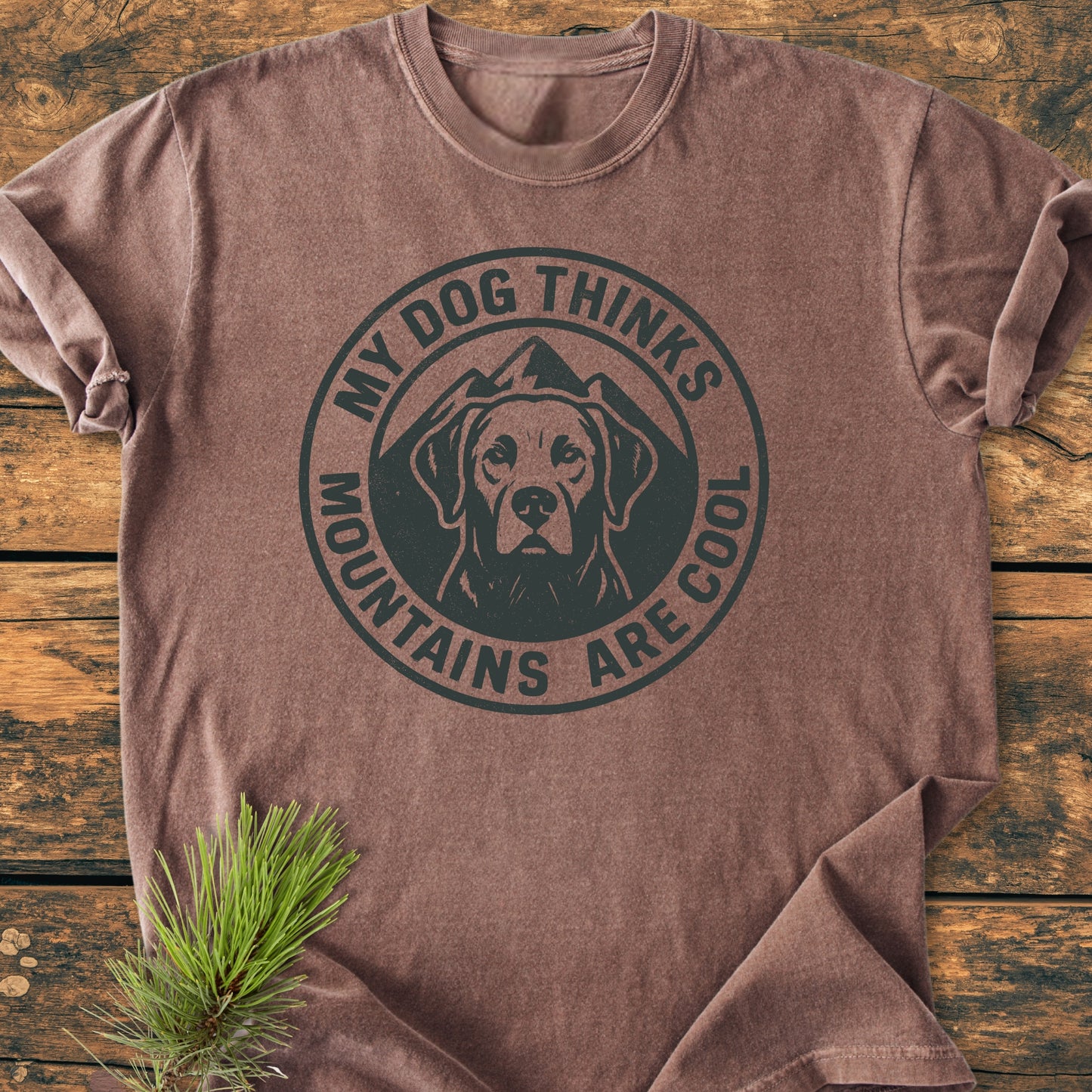 The My Dog Thinks Mountains Are Cool - Vintage Wash Tee features a dog-head graphic with mountain peaks on a faded brown shirt, laid flat with pine needles accentuating its rustic charm.