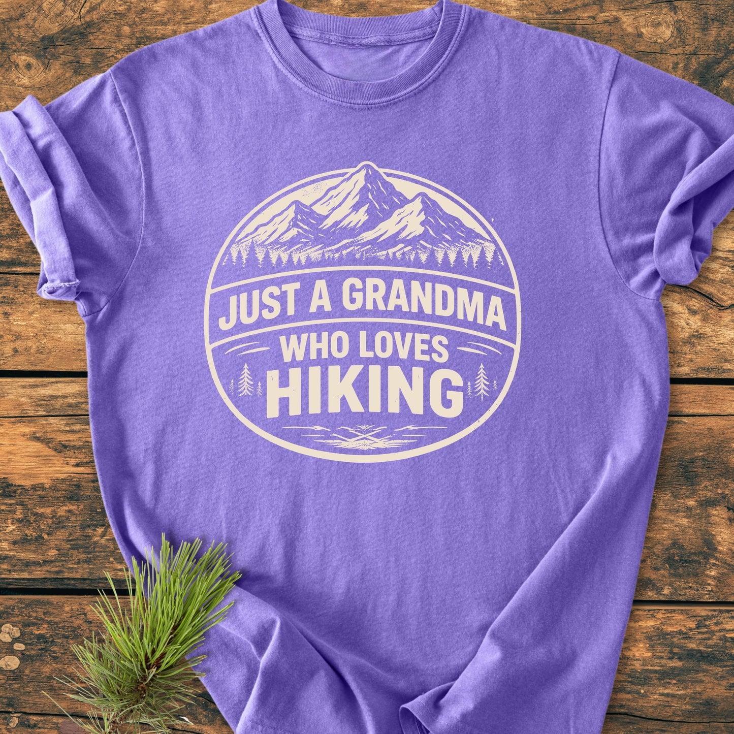 Just A Grandma Who Loves Hiking - Vintage Wash Tee, features a purple shirt with a white circular mountain and pine tree badge, emphasizing comfort and durability with 100% ring-spun cotton and a relaxed fit.
