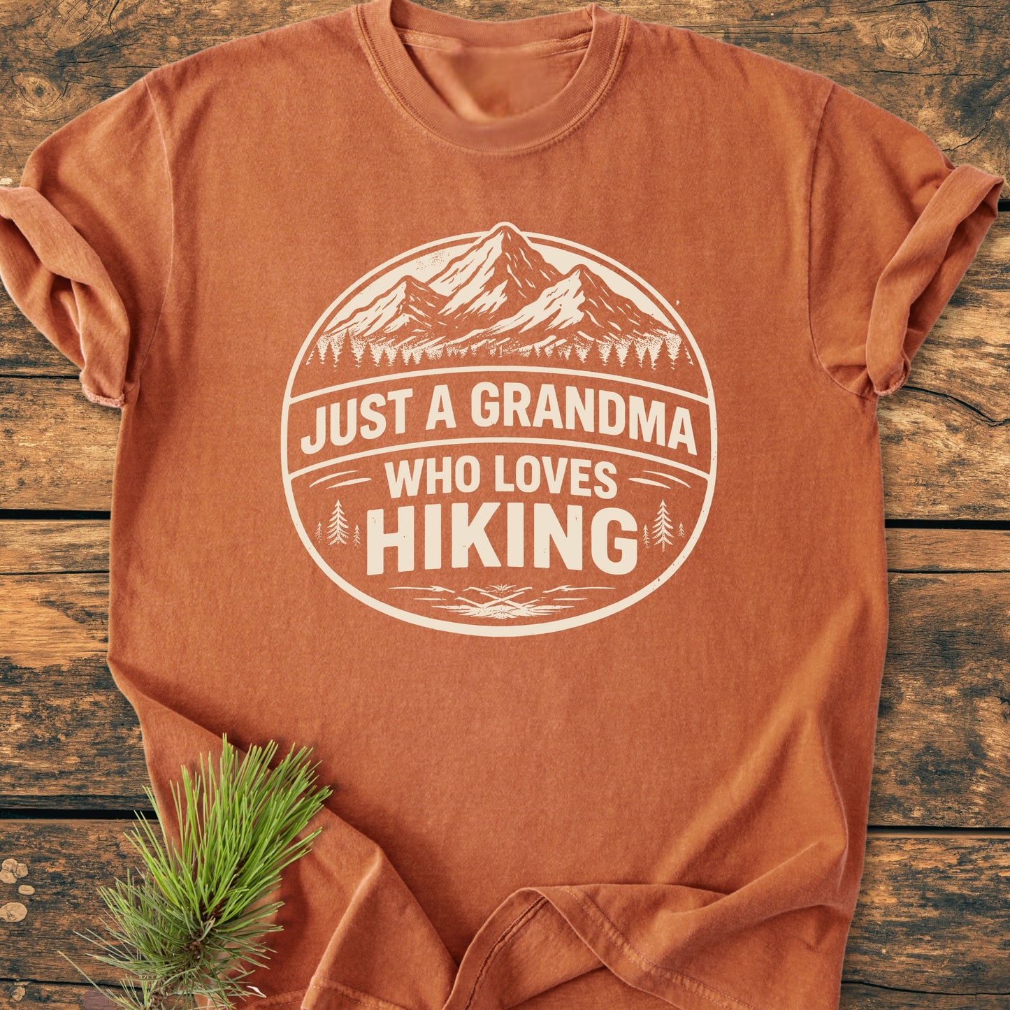 Just A Grandma Who Loves Hiking - Vintage Wash Tee, rust-orange with a mountain and pine tree graphic, laid flat on wood surface, accompanied by a small pine branch, showcasing a cozy, relaxed fit.
