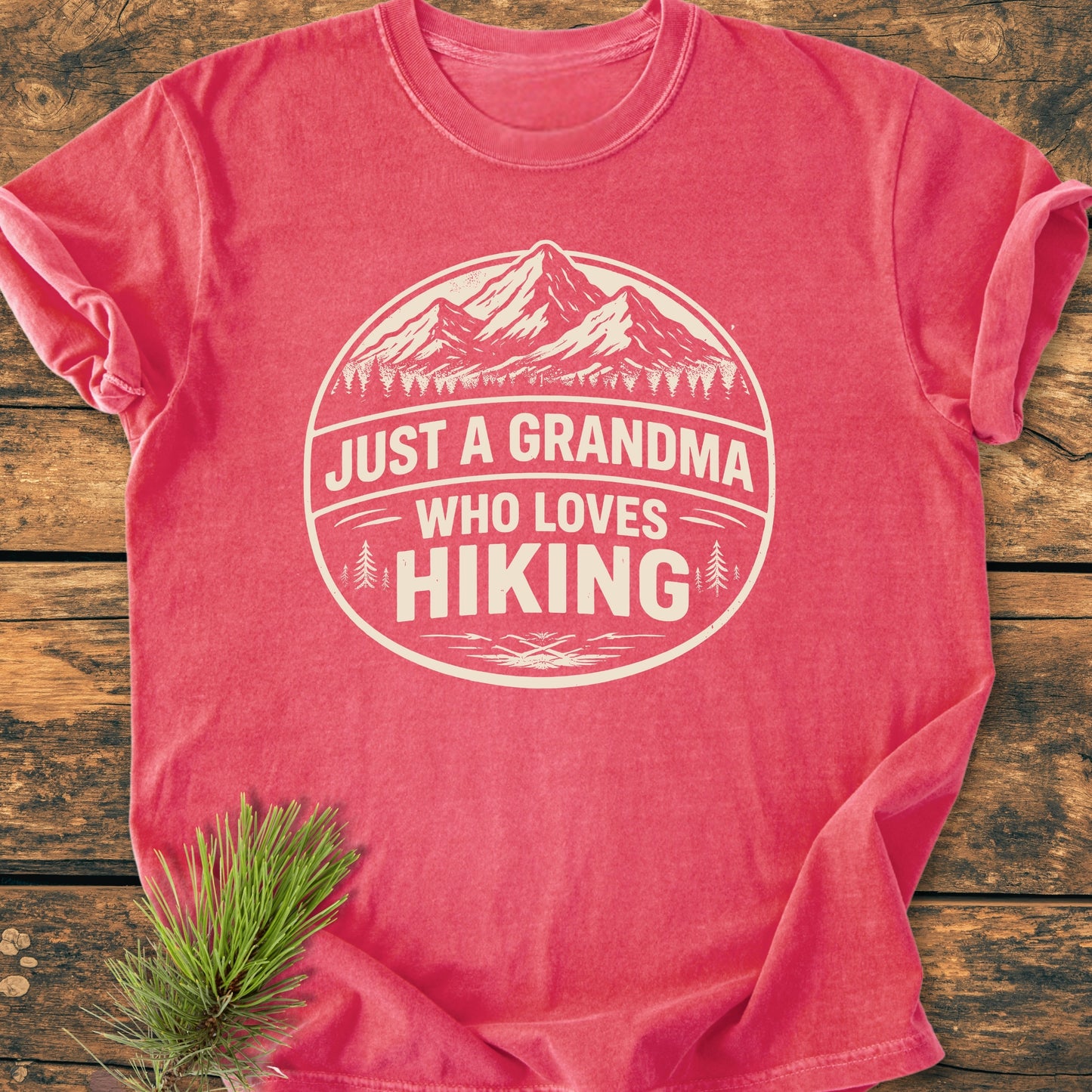 Just A Grandma Who Loves Hiking - Vintage Wash Tee features a cream mountain graphic on a coral-red T-shirt, laid flat with a sprig of evergreen accent, showcasing a cozy, relaxed fit.