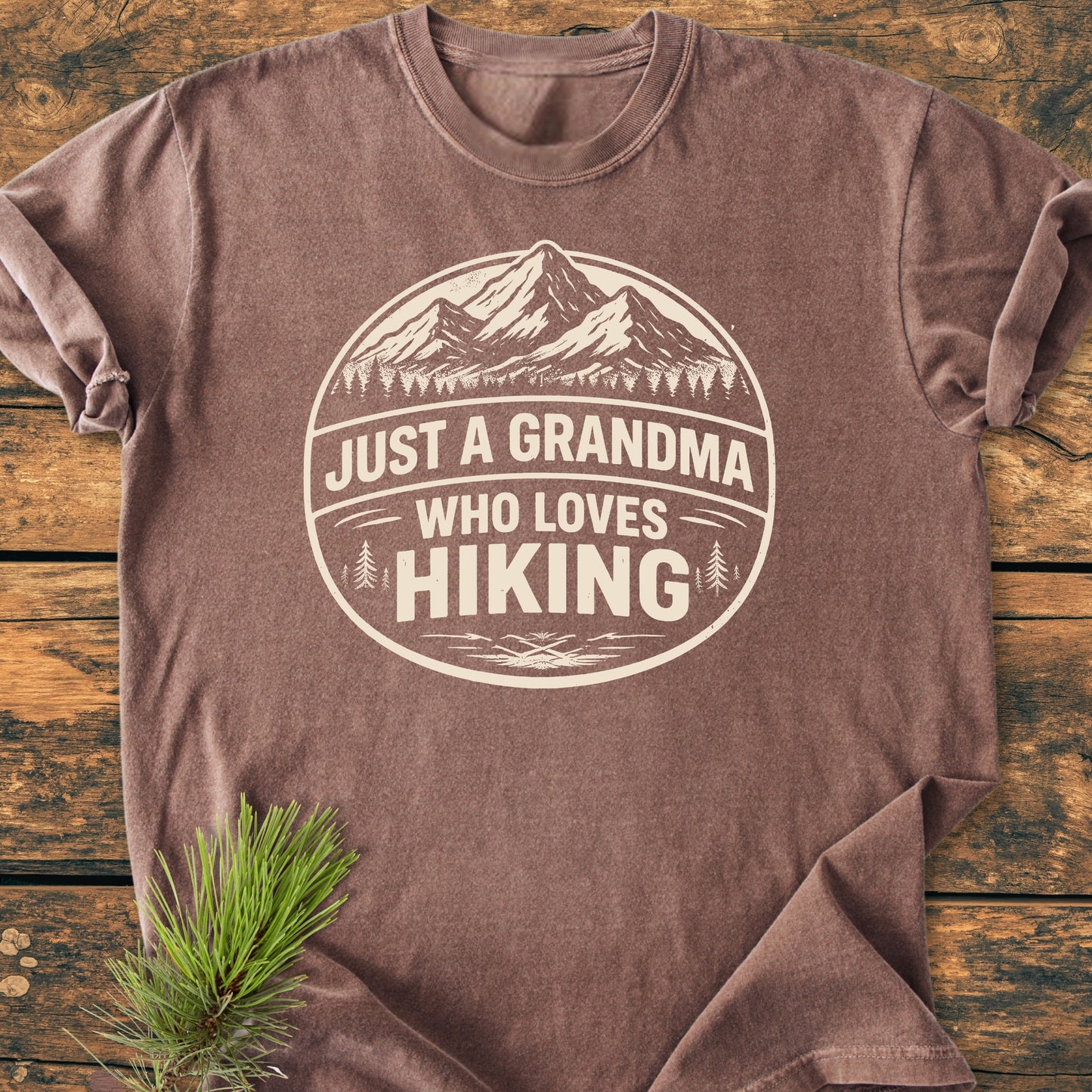 Just A Grandma Who Loves Hiking - Vintage Wash Tee features a circular mountain and tree emblem on a brown, crew-neck design. Laid flat with a pine sprig, highlighting its relaxed fit and durability.