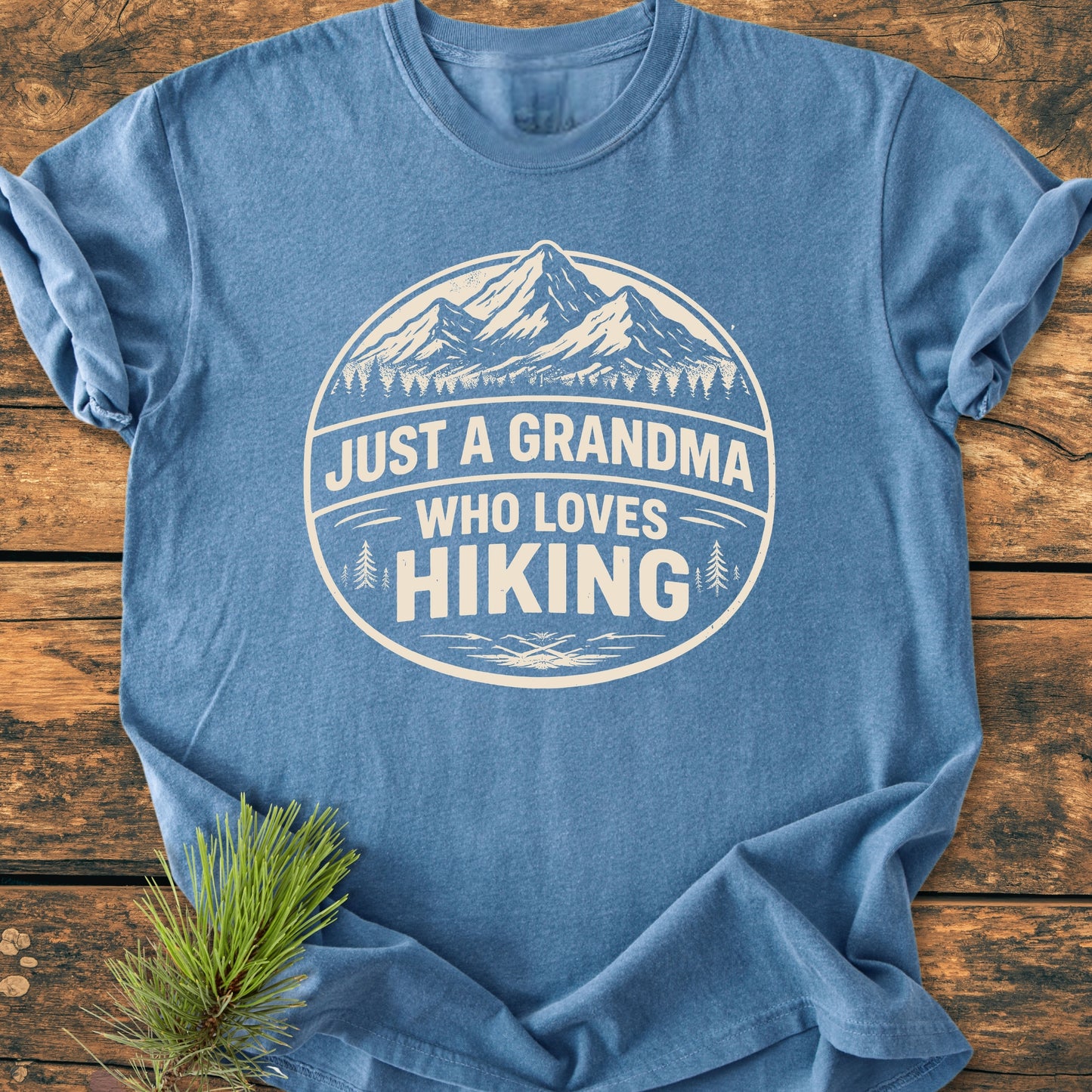 Heather-blue Just A Grandma Who Loves Hiking - Vintage Wash Tee with a mountain range and pine trees graphic, laid on wood, showcasing its relaxed fit and durable, soft-washed fabric.