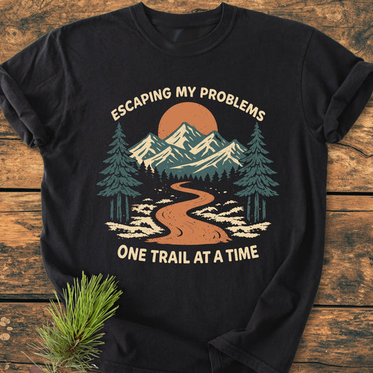 Escaping My Problems - Vintage Wash Tee features a stylized outdoor scene with trail, forest, mountains, and sun, on a dark t-shirt, displayed with a pine sprig on a wooden surface.
