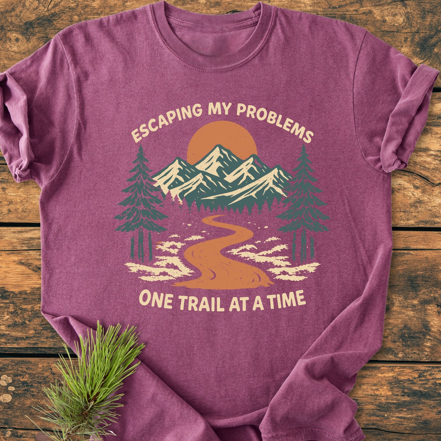 Heathered purple Escaping My Problems - Vintage Wash Tee features a stylized outdoor scene with pine trees, mountain range, and sun/moon. Laid flat with a pine branch beside it on wooden surface.