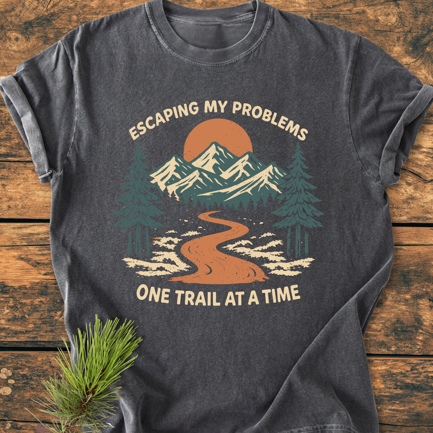 Escaping My Problems - Vintage Wash Tee features a vintage outdoors graphic with mountains, a trail, and an orange sun, on a soft, dark-gray T-shirt laid flat on wood.